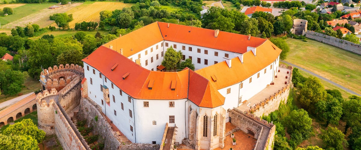 A historic castle with a red-tiled roof, surrounded by lush greenery and a stone wall.