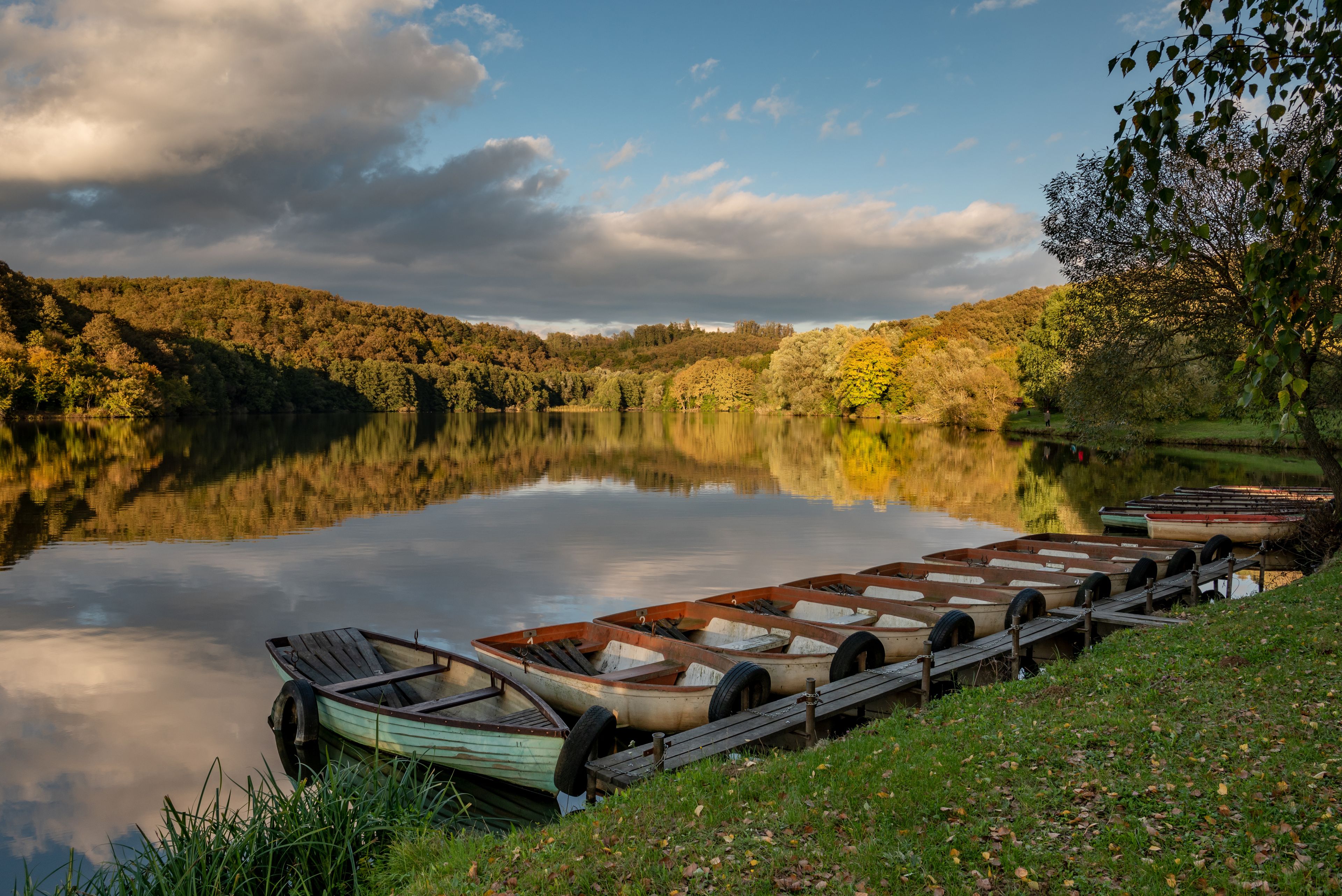 A calm lake with boats docked on the shore, surrounded by autumn foliage and a cloudy sky.