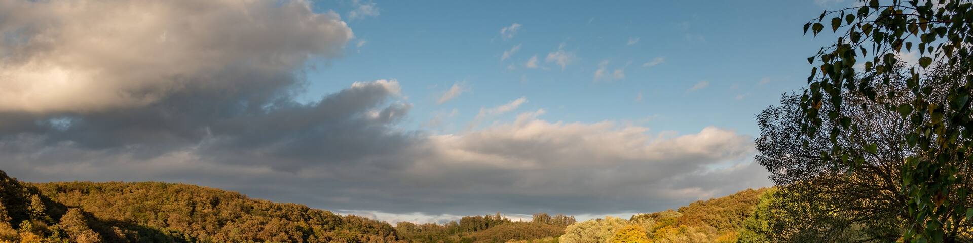A calm lake with boats docked on the shore, surrounded by autumn foliage and a cloudy sky.