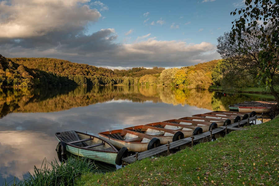 A calm lake with boats docked on the shore, surrounded by autumn foliage and a cloudy sky.