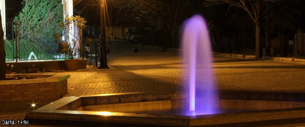 A nighttime scene with a fountain, a cross atop a structure, and a street lamp.