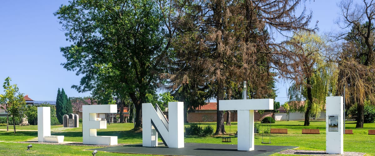 A park with large white letters spelling "LENTI" on a grassy area, surrounded by trees and benches.