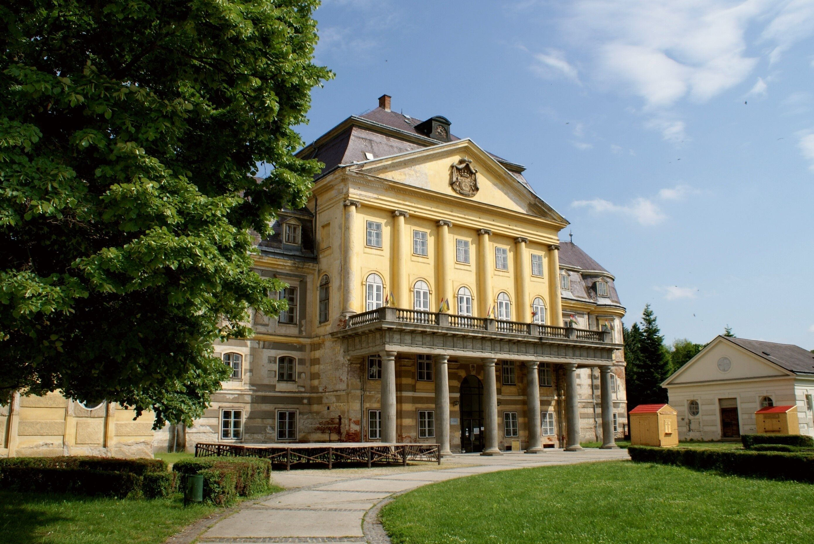 A large yellow building with columns and a balcony, surrounded by greenery.
