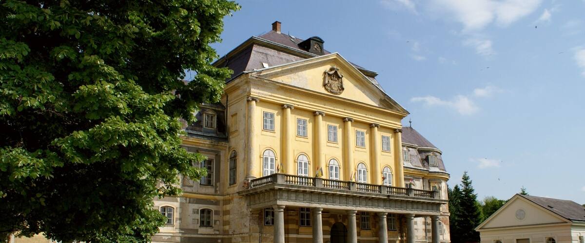 A large yellow building with columns and a balcony, surrounded by greenery.