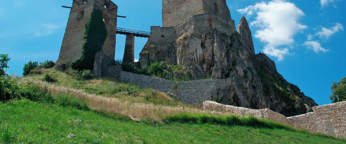 A historic stone castle with a bridge, surrounded by lush green hills and a clear blue sky.