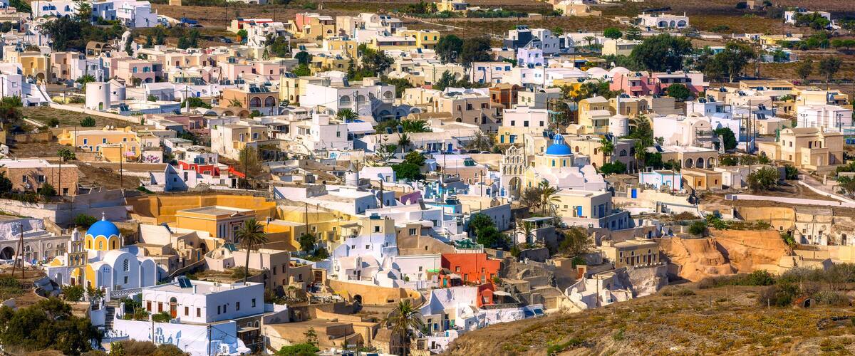 A densely packed coastal town with white and pastel-colored buildings, some with blue domes, set against a hilly backdrop.