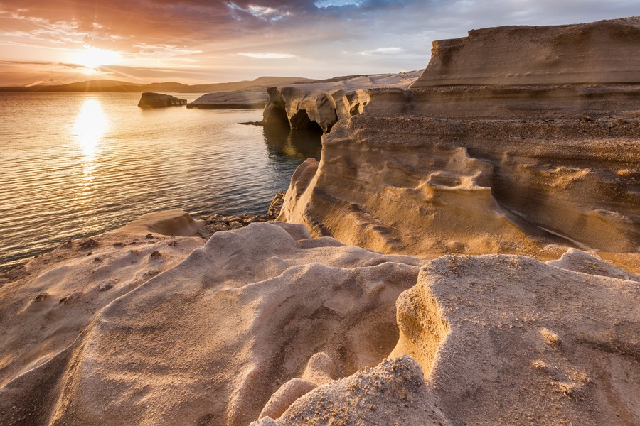 A coastal landscape with layered rock formations and a calm sea at sunset.