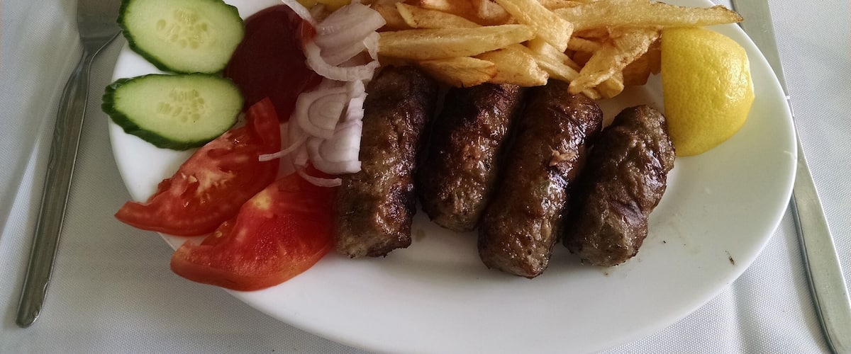 A plate with sausages, fries, lemon, cucumber slices, tomato, and onion rings.