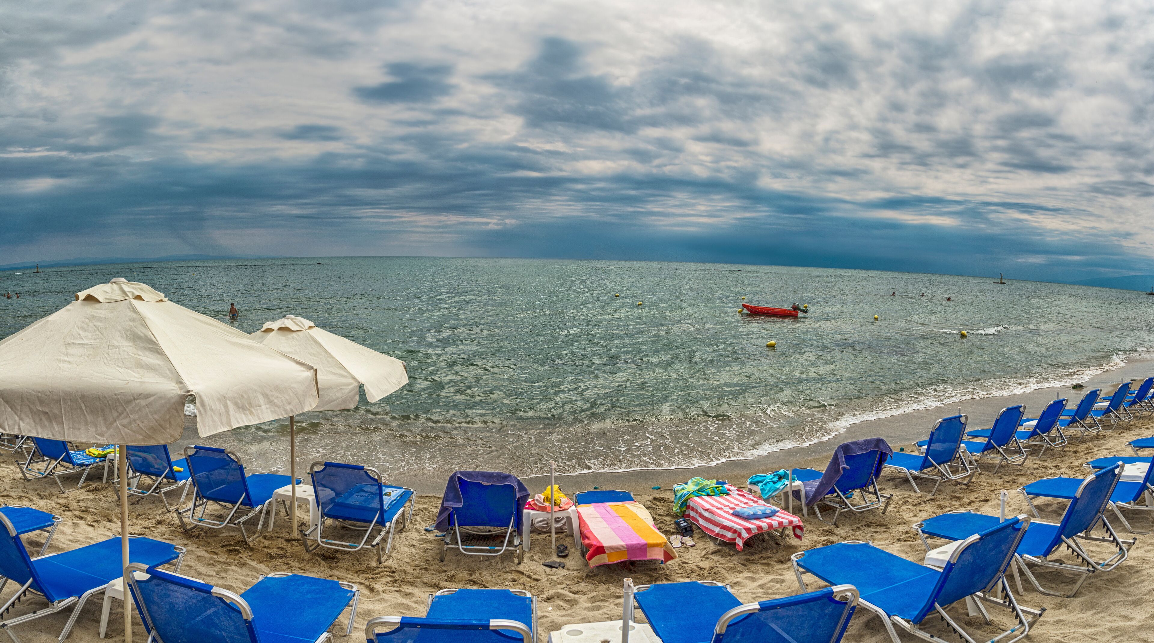 A beach with blue chairs and white umbrellas, a red boat in the water, and a cloudy sky.