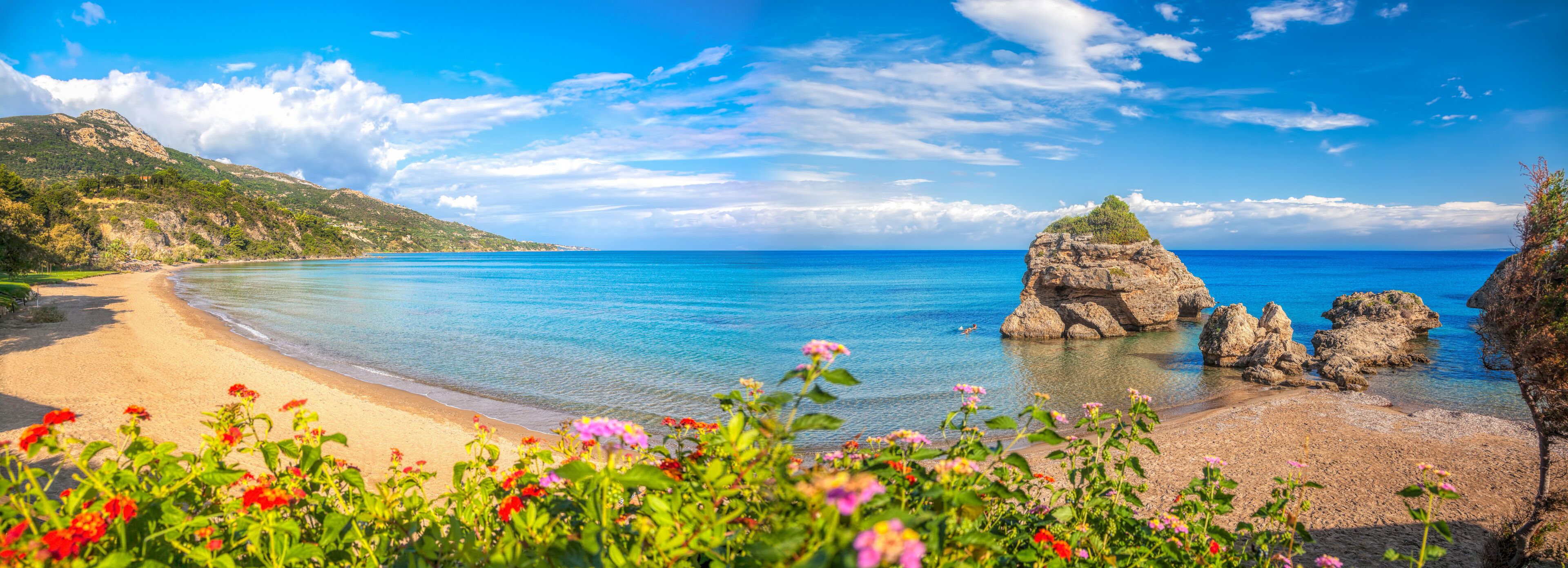 A beach with clear blue water, a rocky outcrop, and a mountainous backdrop.