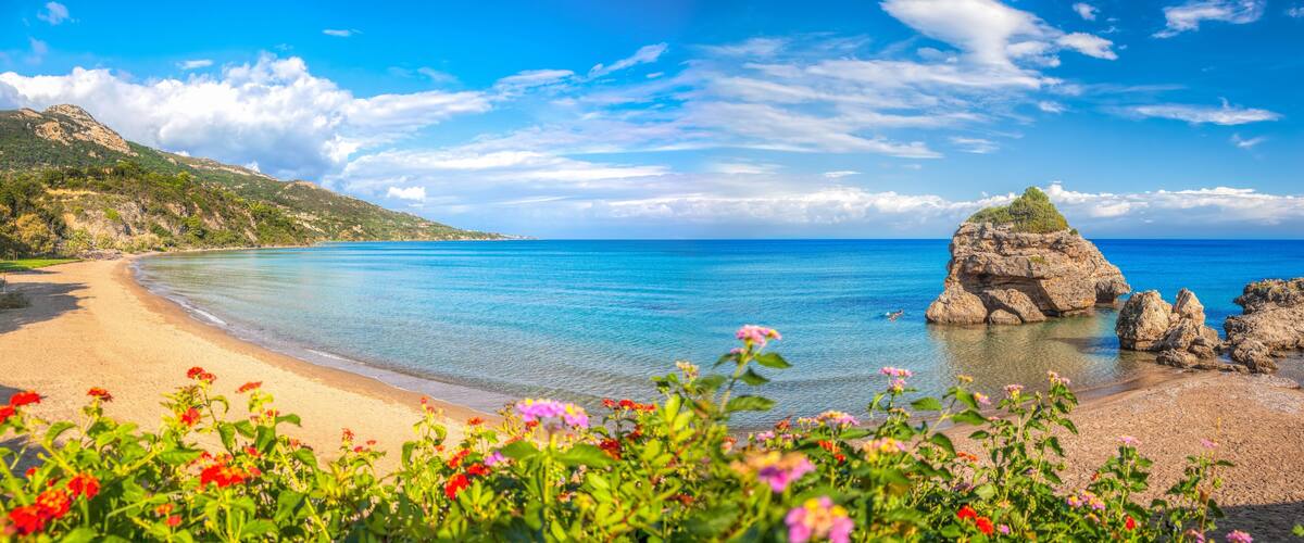 A beach with clear blue water, a rocky outcrop, and a mountainous backdrop.