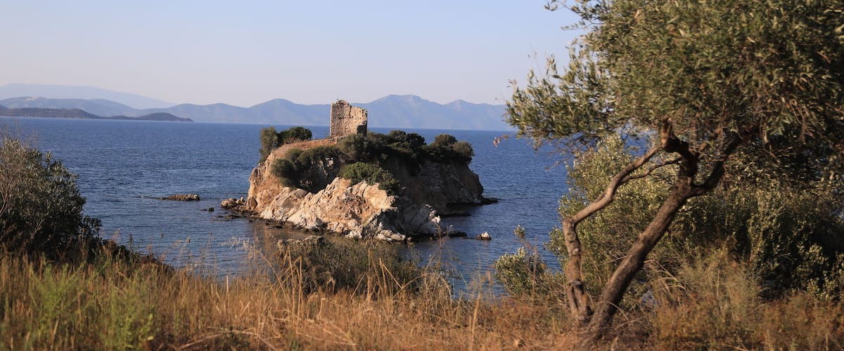 Un paysage côtier avec une île rocheuse sur laquelle se dresse une petite tour, le tout entouré d’une eau d’un bleu limpide et dominé par des montagnes lointaines.
