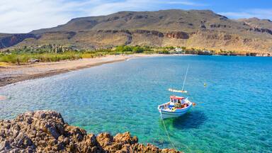 A boat anchored near a rocky shoreline with clear blue water and a mountainous backdrop.