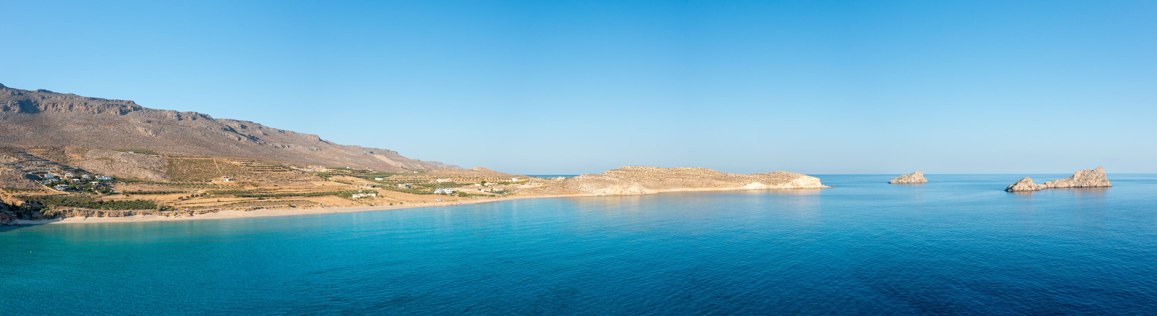 A coastal landscape with a clear blue sea, rocky outcrops, and a small settlement with buildings near the shore.