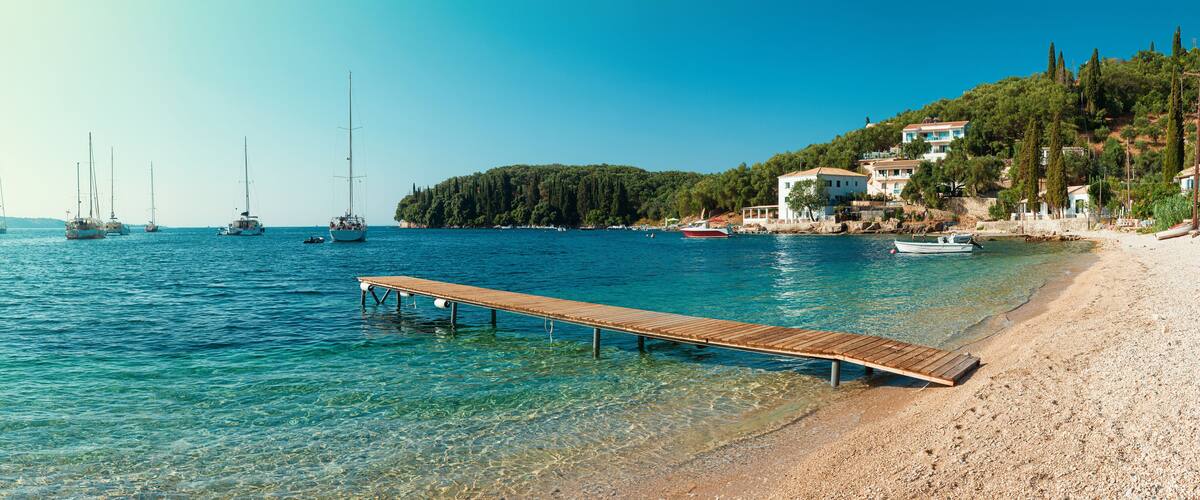 A coastal scene with a wooden pier extending into clear blue waters, boats anchored nearby, and buildings on a hillside.