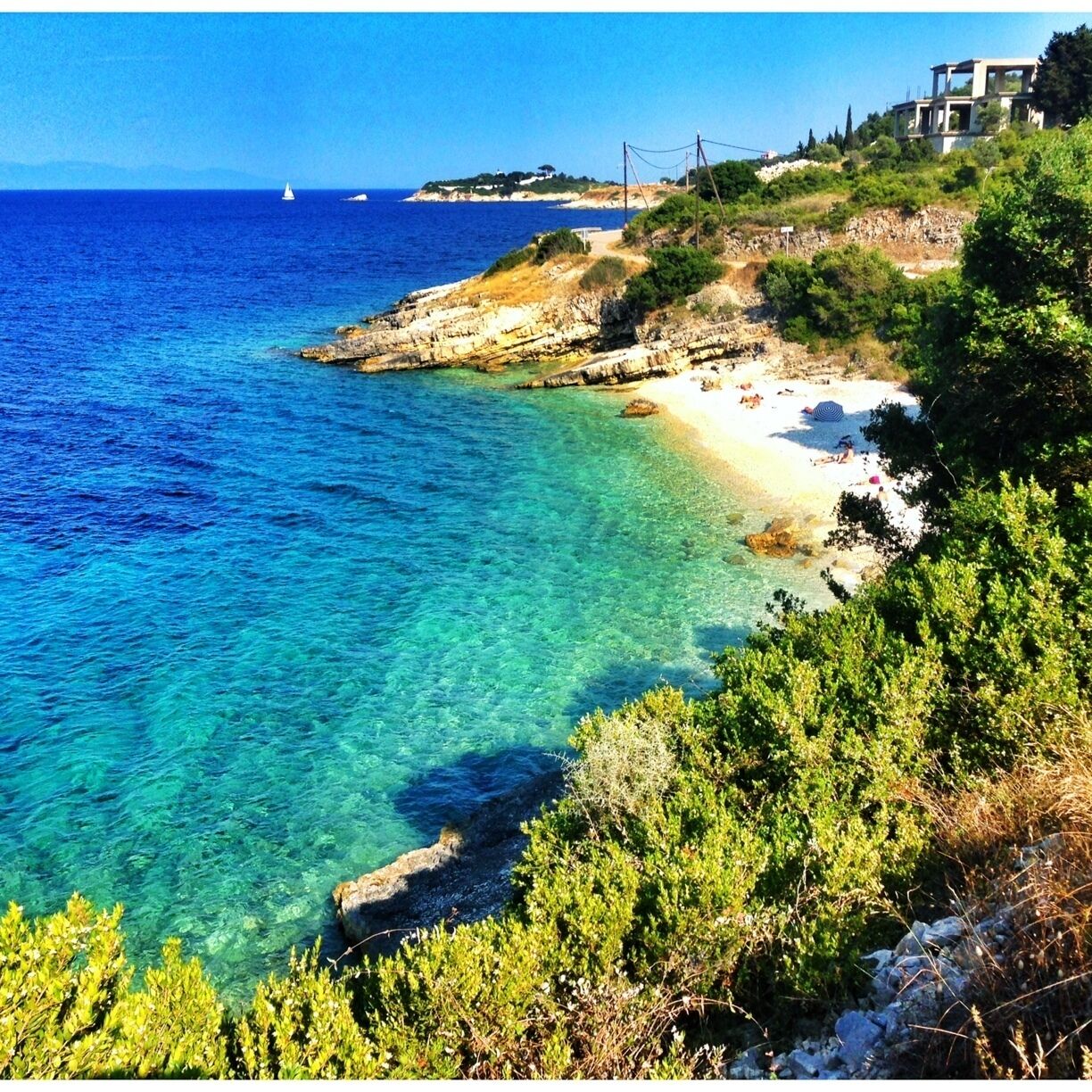 A coastal landscape with clear turquoise waters, a sandy beach, rocky cliffs, and a modern house on a hillside.