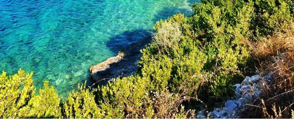 A coastal landscape with clear turquoise waters, a sandy beach, rocky cliffs, and a modern house on a hillside.