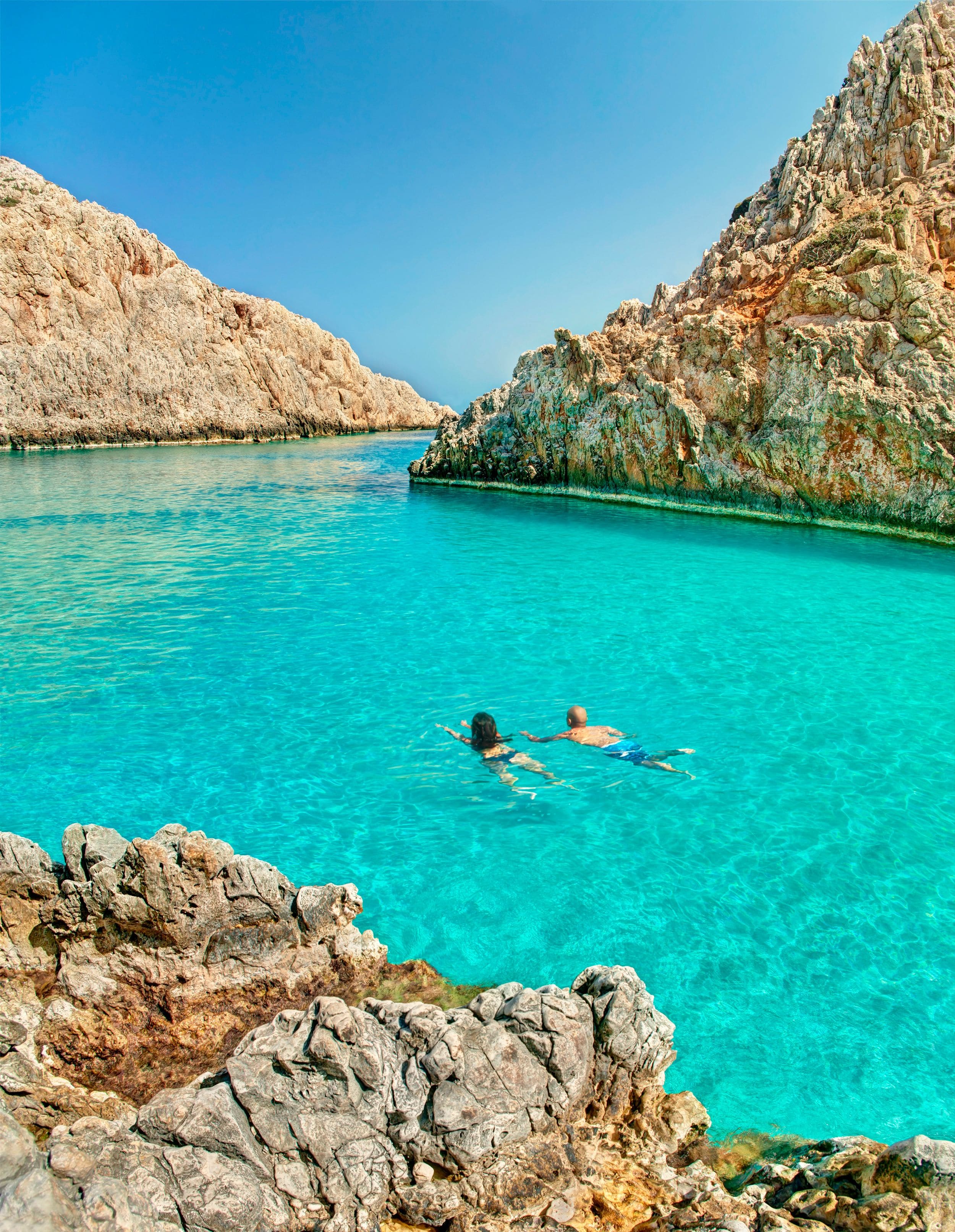 A clear turquoise sea with rocky cliffs and two people swimming.