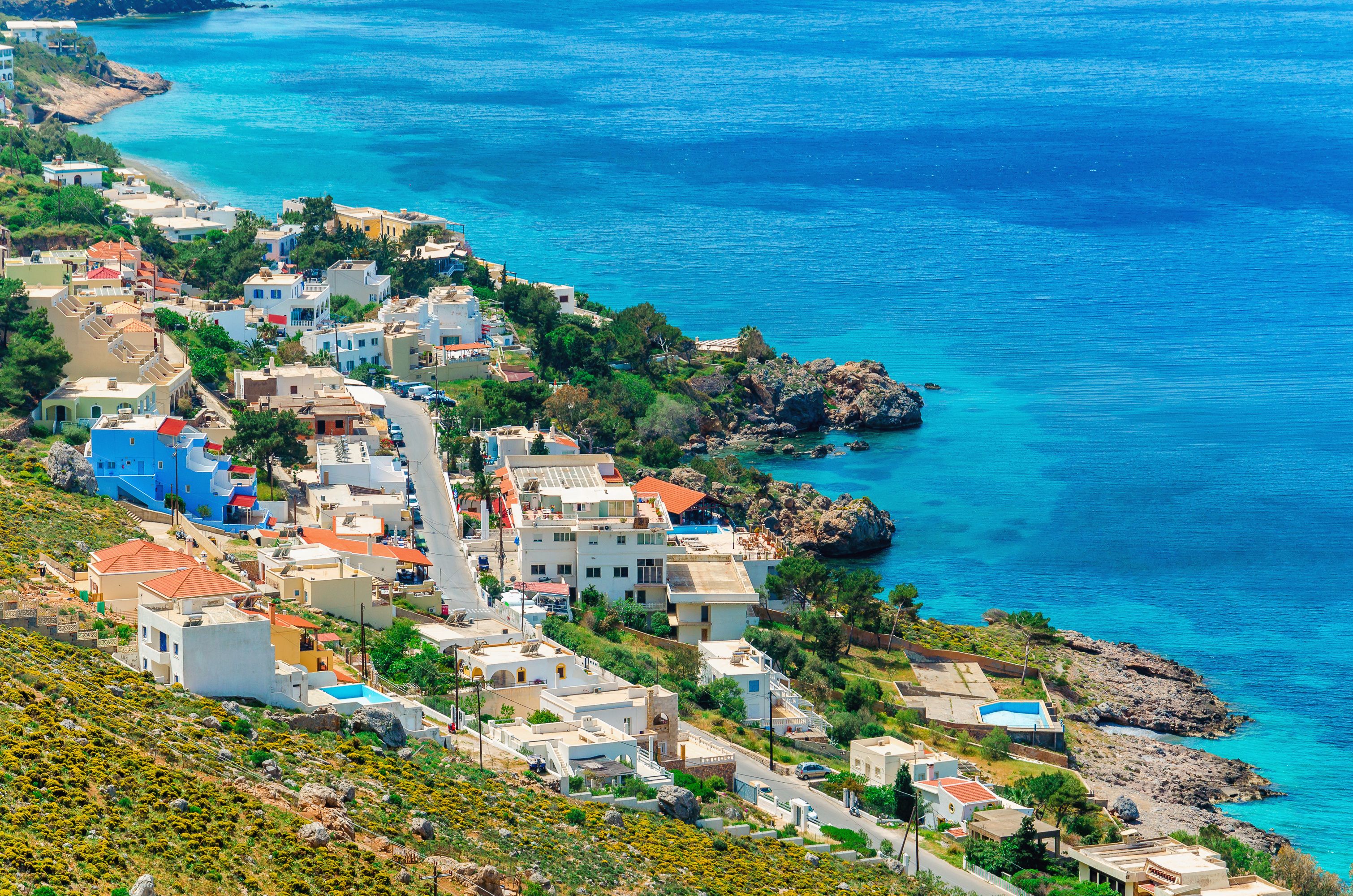 A coastal town with buildings along the shoreline, a rocky outcrop, and clear blue waters.