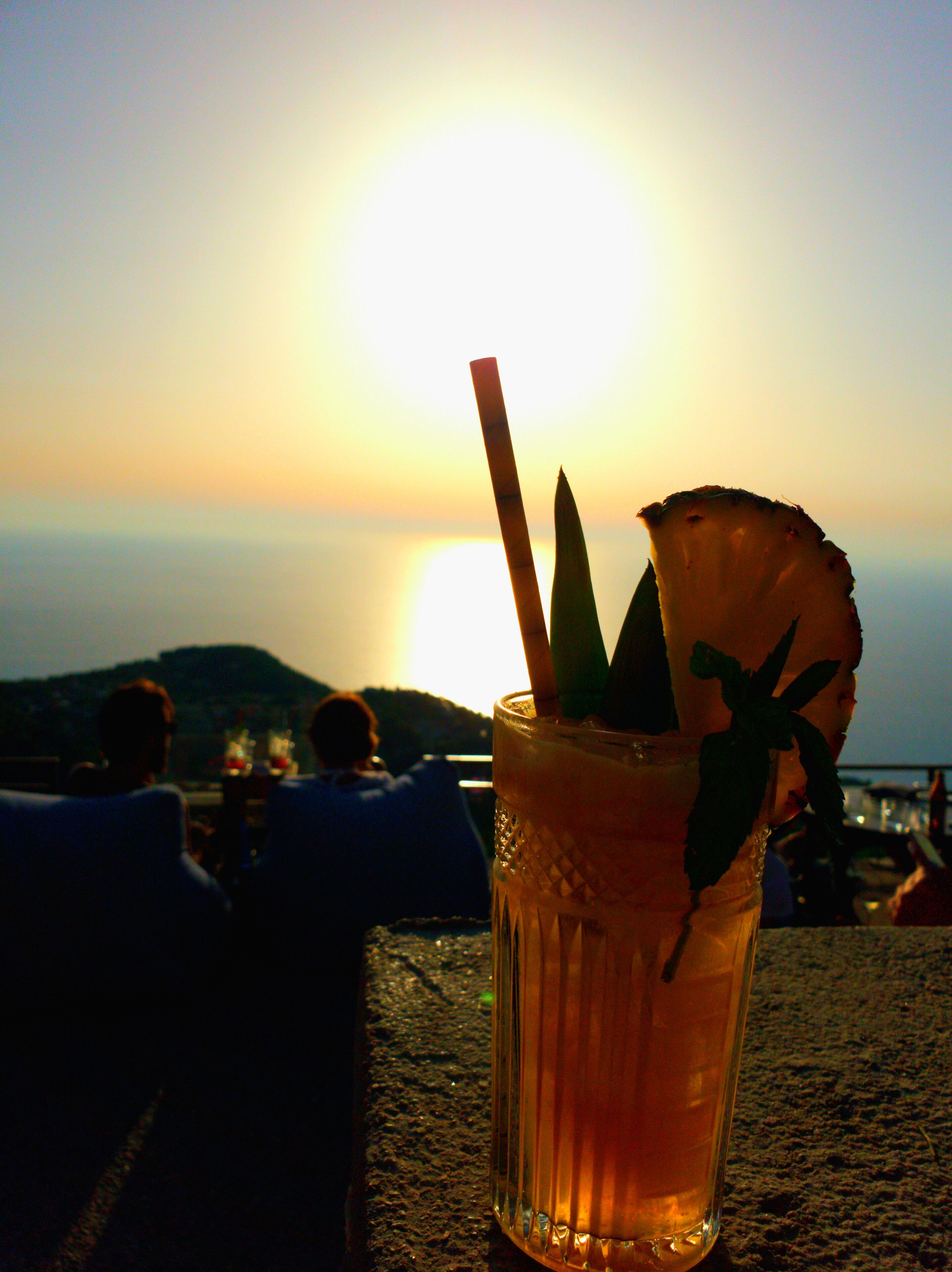 A cocktail glass with a straw and a slice of pineapple on a table with a sunset in the background.
