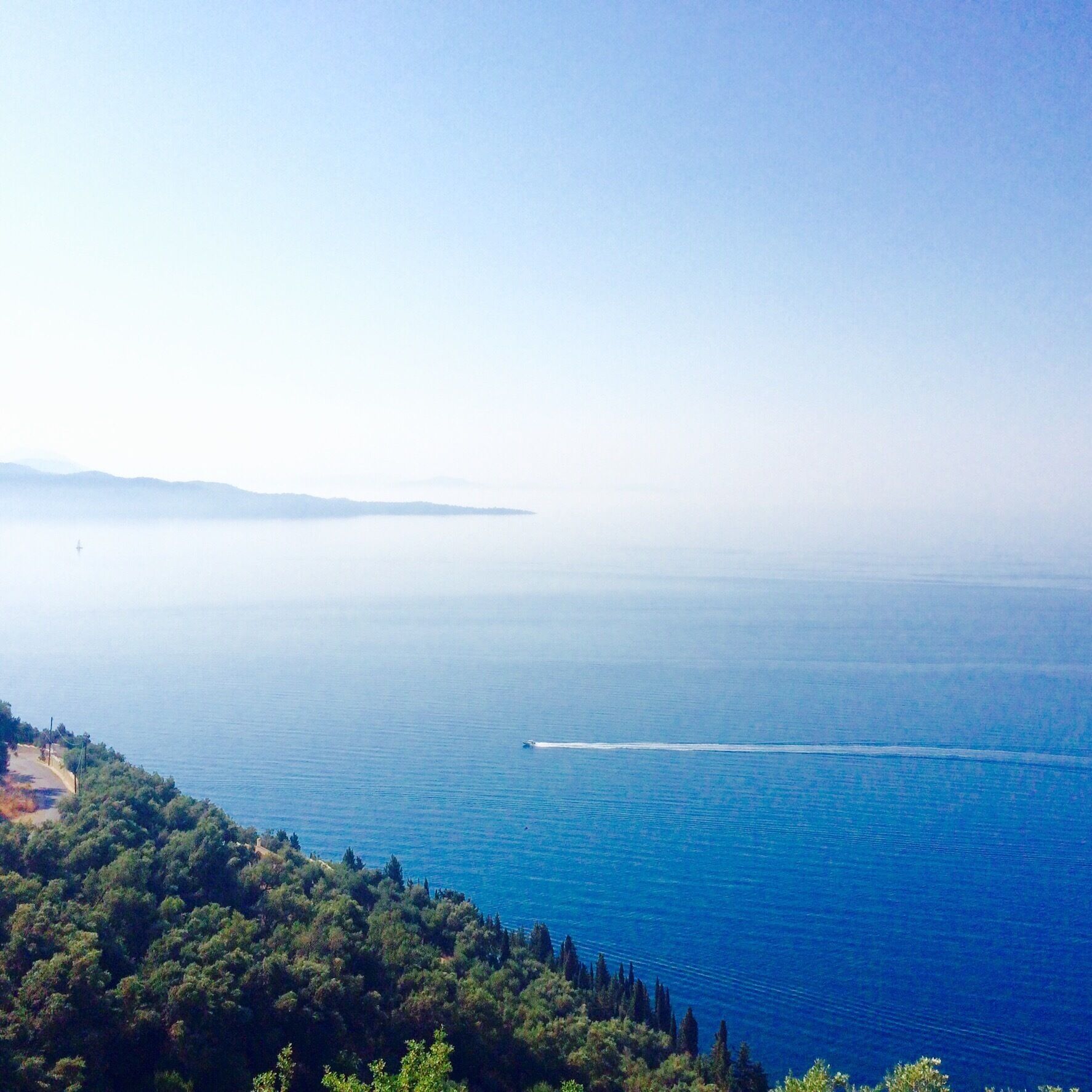 A coastal landscape with a clear view of the sea, a distant shoreline, and a road winding through a forested area.