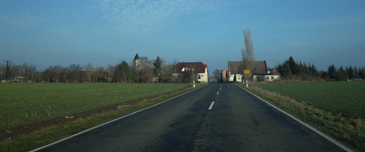 A rural road flanked by fields and houses, leading to a church with a steeple.