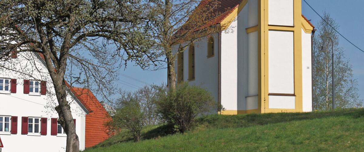 A church with a tall steeple, a white building with red shutters, and a tree in the foreground.