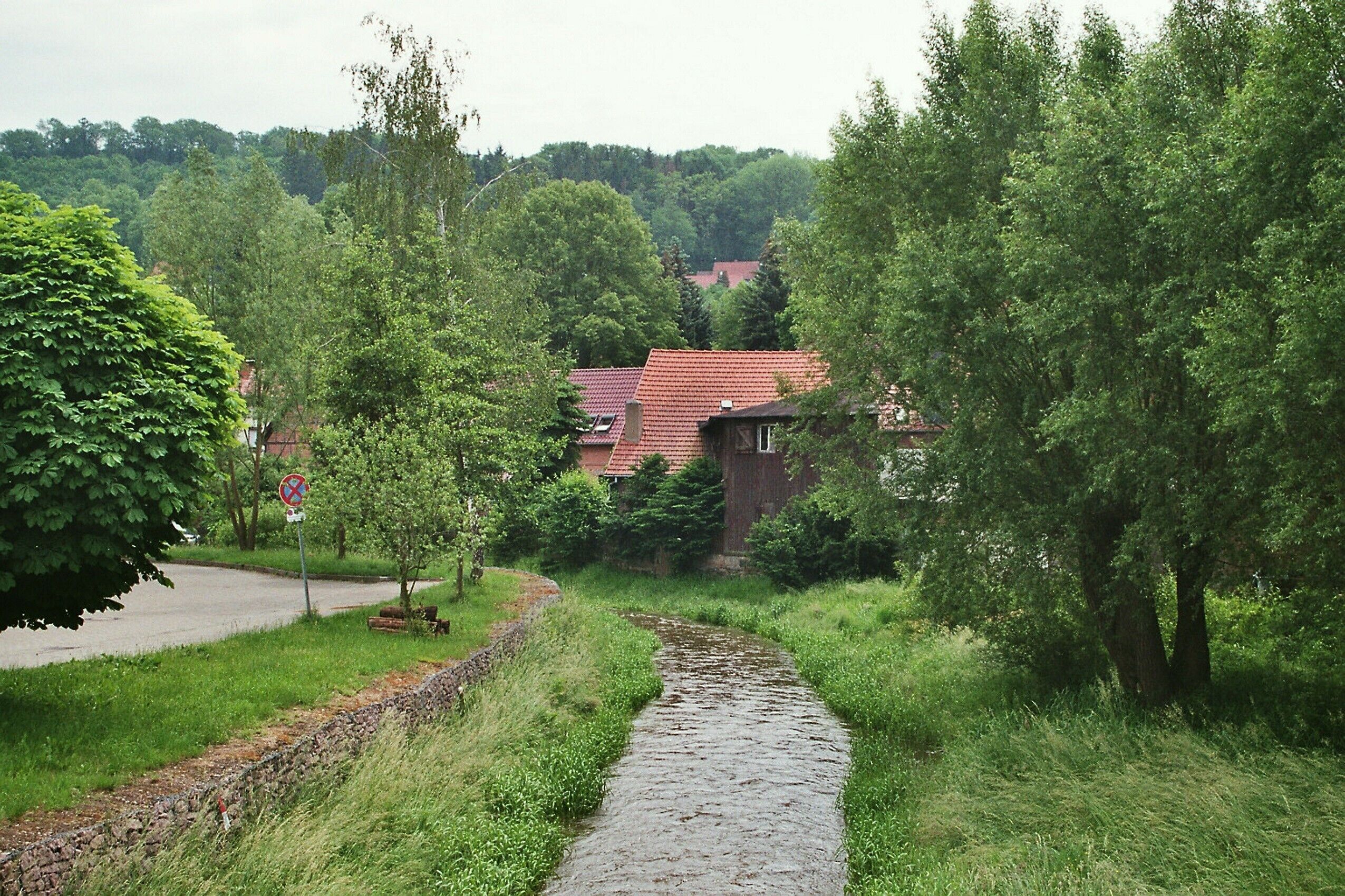 A narrow stone path winding through a lush green landscape with trees and a red-roofed building in the background.