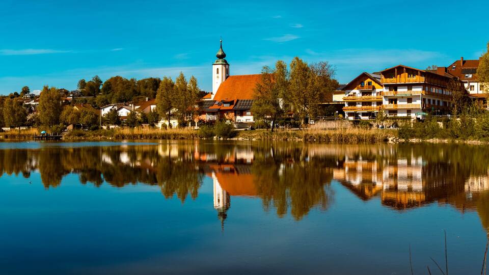 A calm lake reflecting buildings and trees, with a church steeple and modern houses along the shore.