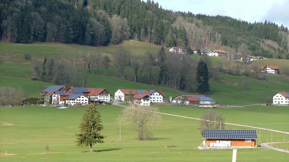 A rural landscape with a cluster of houses on a hillside, surrounded by green fields and a forested mountain.