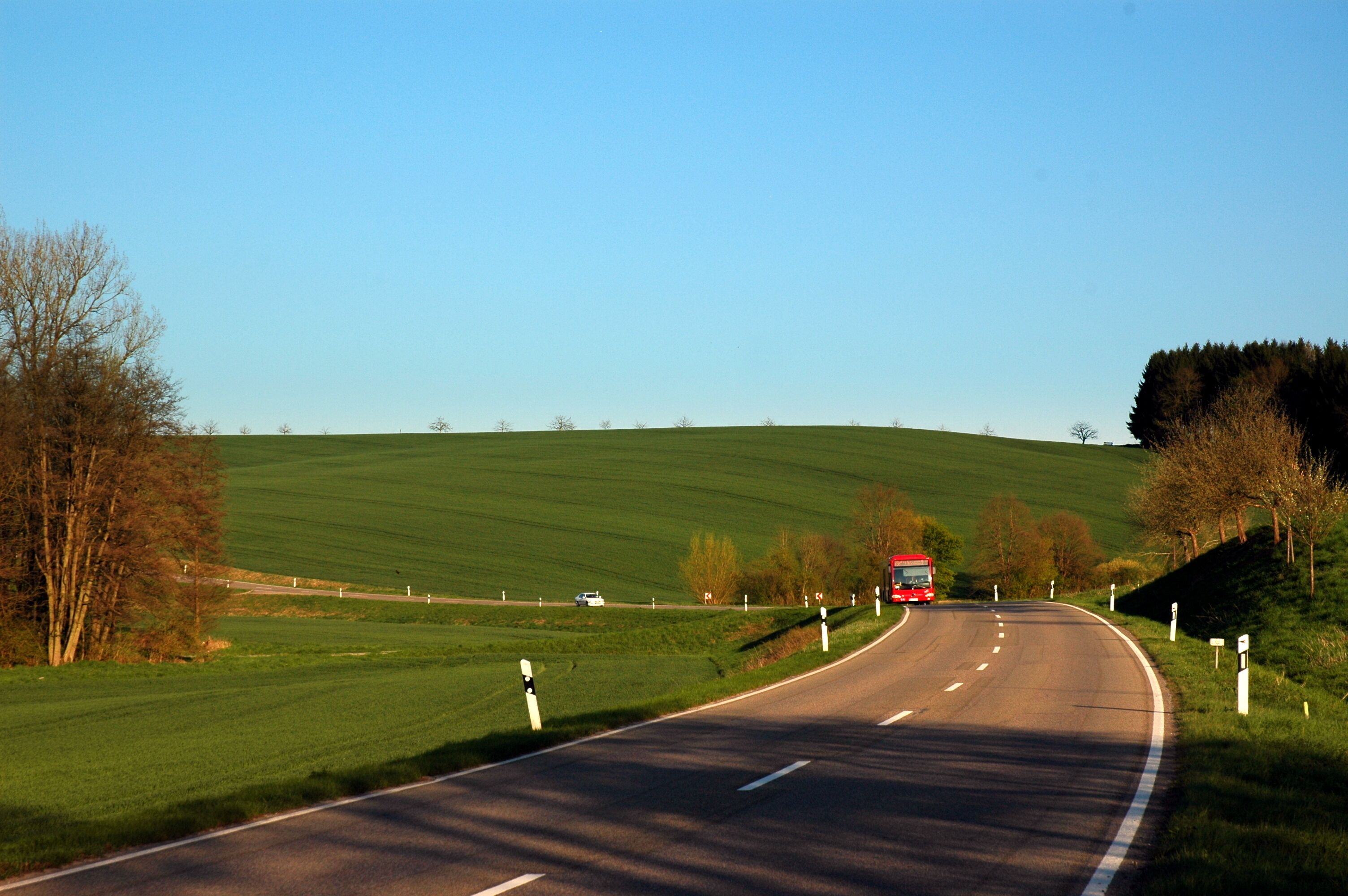 A rural road curving through a green landscape with a red bus approaching.