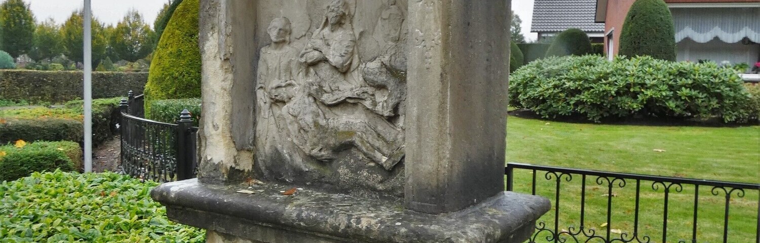 Un monumento de piedra con un relieve escultórico, rodeado de setos cuidados y un muro de ladrillo.
