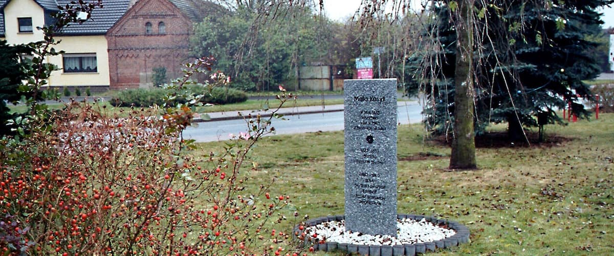 A stone monument surrounded by a circular gravel path, with a house and a tree in the background.