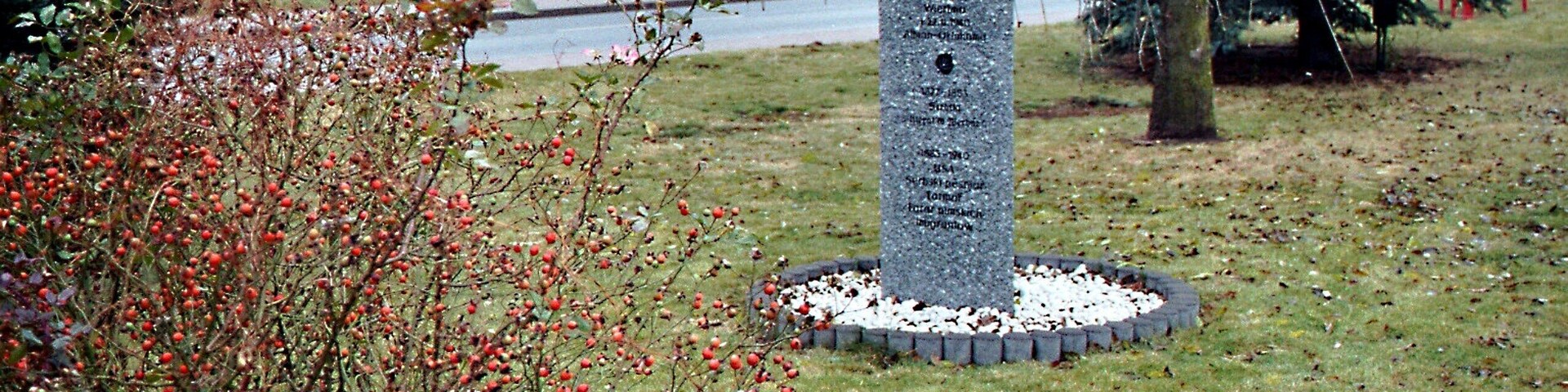 A stone monument surrounded by a circular gravel path, with a house and a tree in the background.