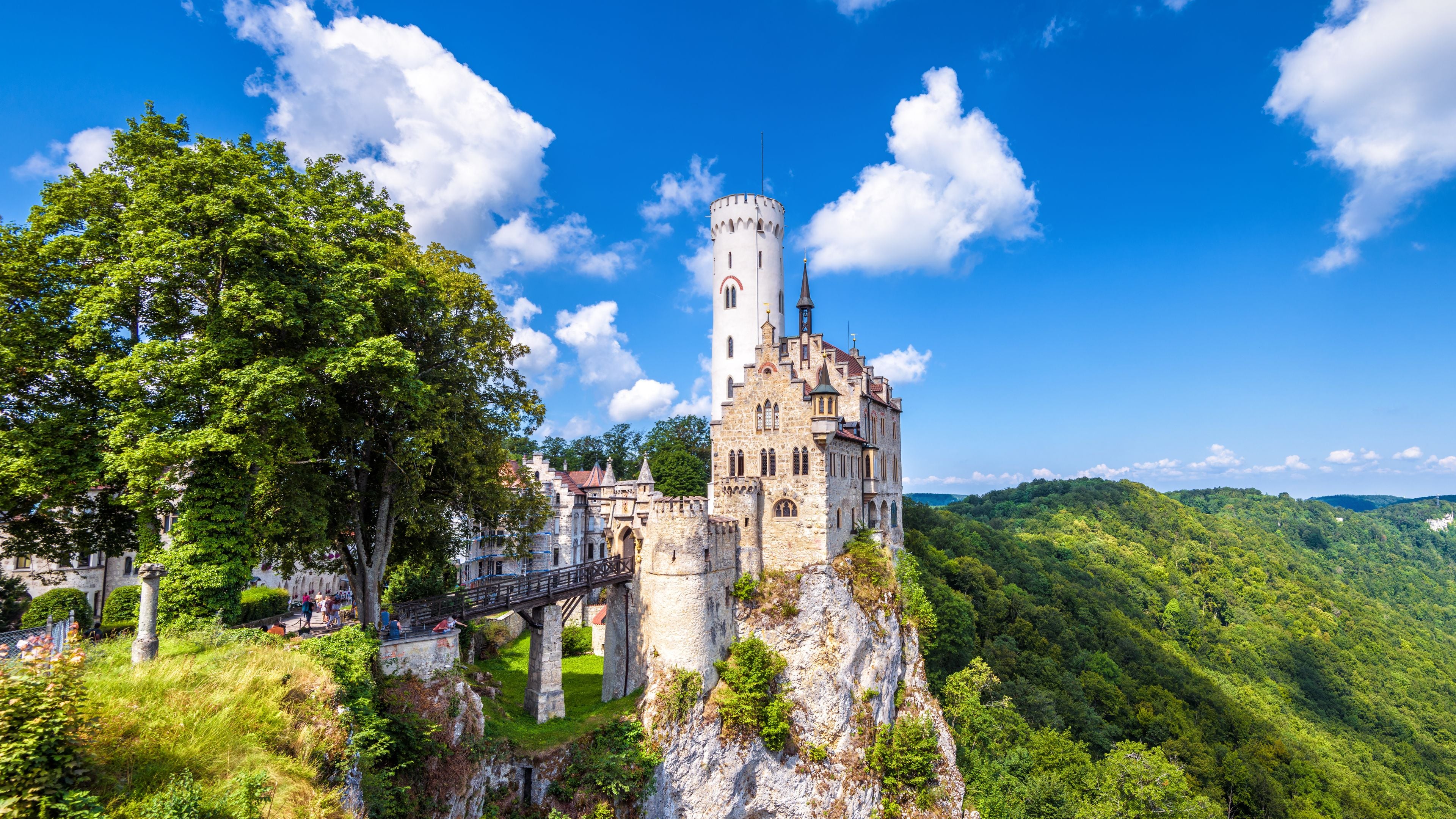 A castle on a cliff with a bridge, surrounded by lush greenery and a clear blue sky.