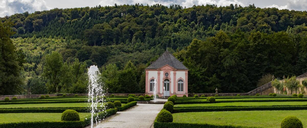A formal garden with a fountain, trimmed hedges, and a building with a clock tower.
