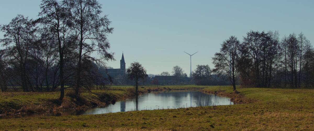 A serene landscape with a pond, trees, a church steeple, and wind turbines in the distance.