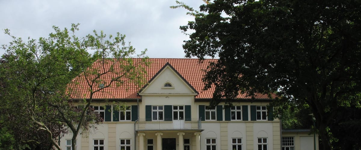 A large, two-story building with a red-tiled roof and green shutters.
