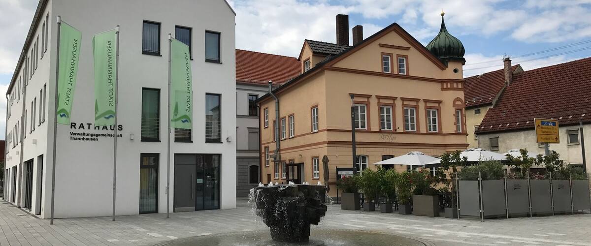 A public square with a fountain, a white building with green banners, and a traditional building with a green dome.