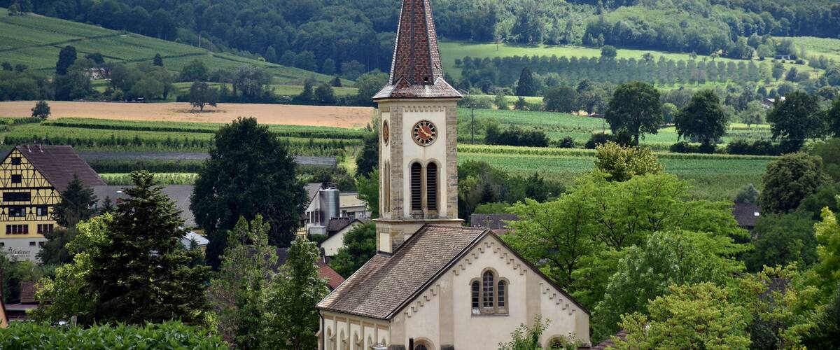A church with a tall steeple, surrounded by lush green fields and a forested hill in the background.
