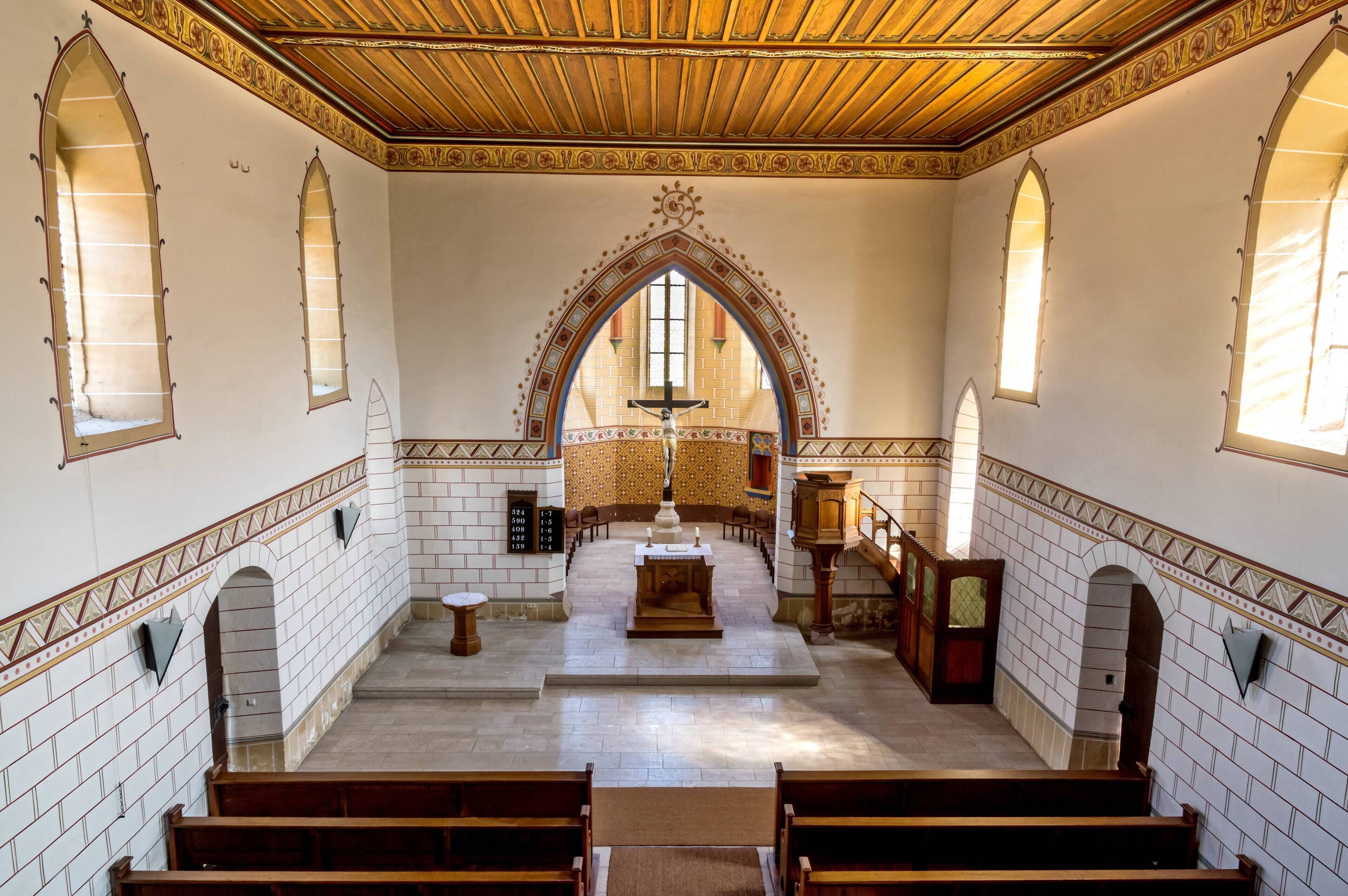 A church interior with arched windows, a wooden ceiling, and a crucifix at the altar.