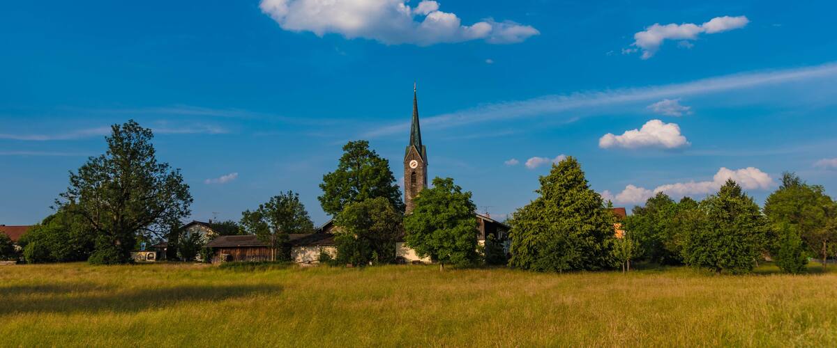 A church with a tall steeple, surrounded by trees and open fields under a clear blue sky.