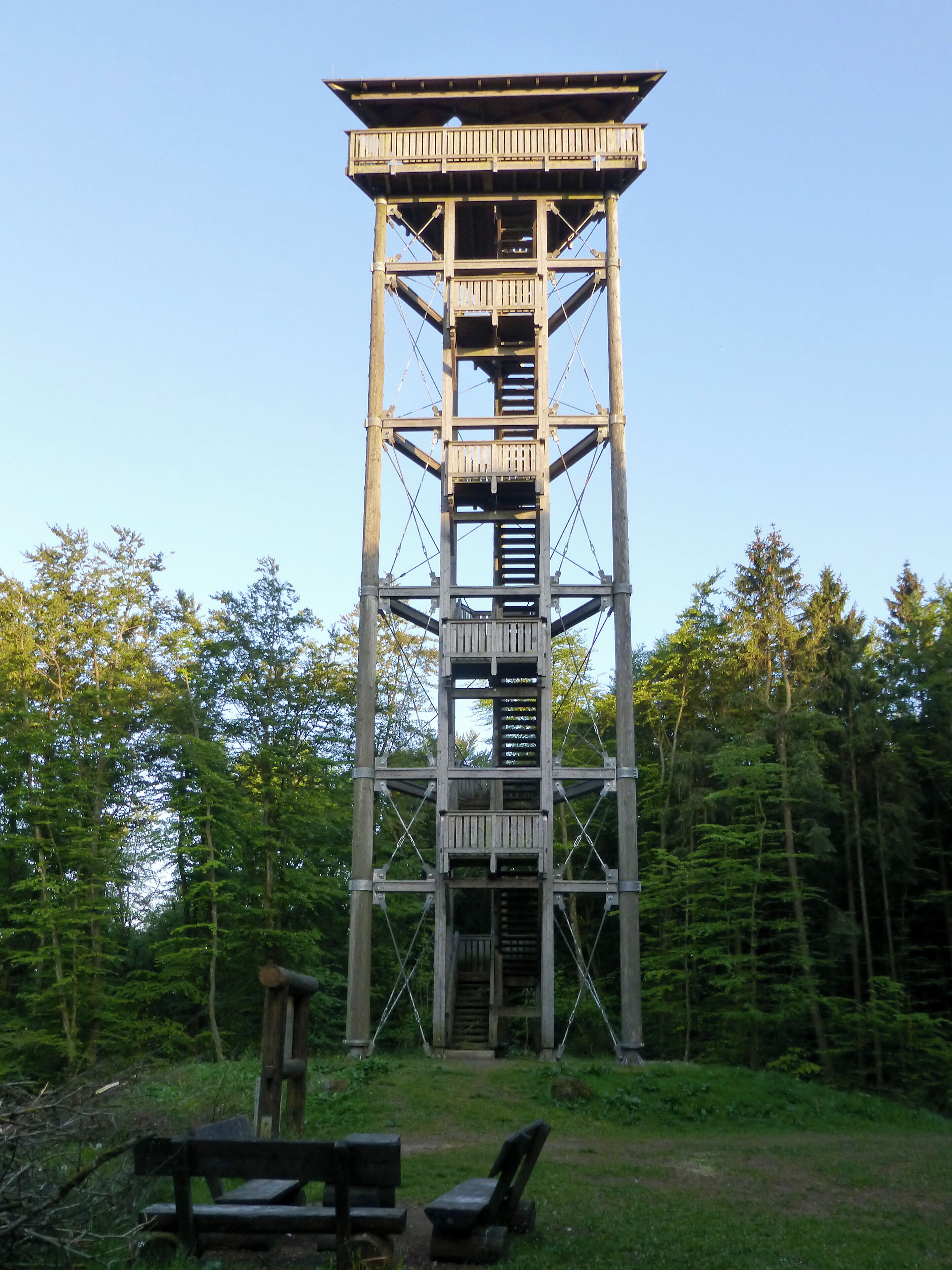 A tall wooden tower with multiple platforms and railings, surrounded by a forested area with benches at the base.