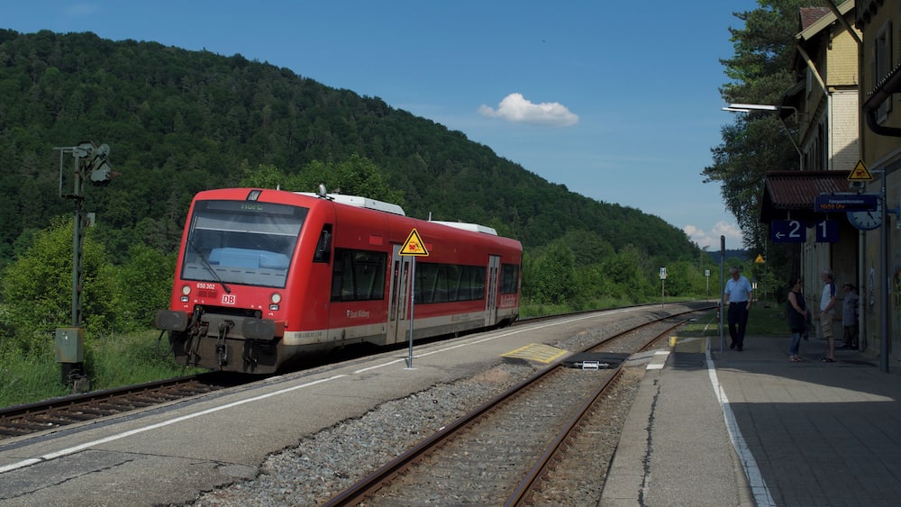 Un train rouge est garé dans une gare, avec une colline boisée en arrière-plan.