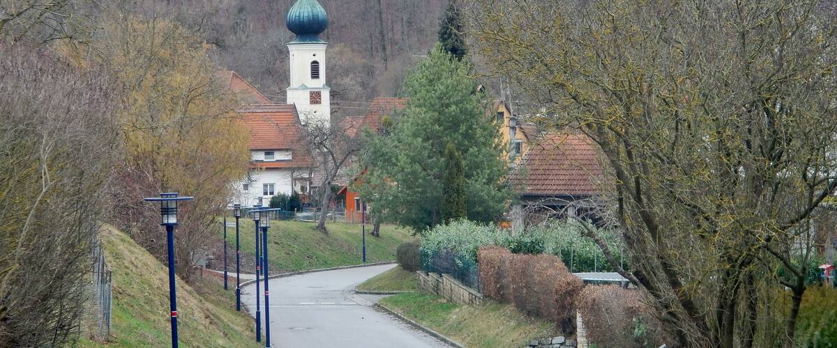 A village with a church featuring a green dome, surrounded by trees and a winding road.