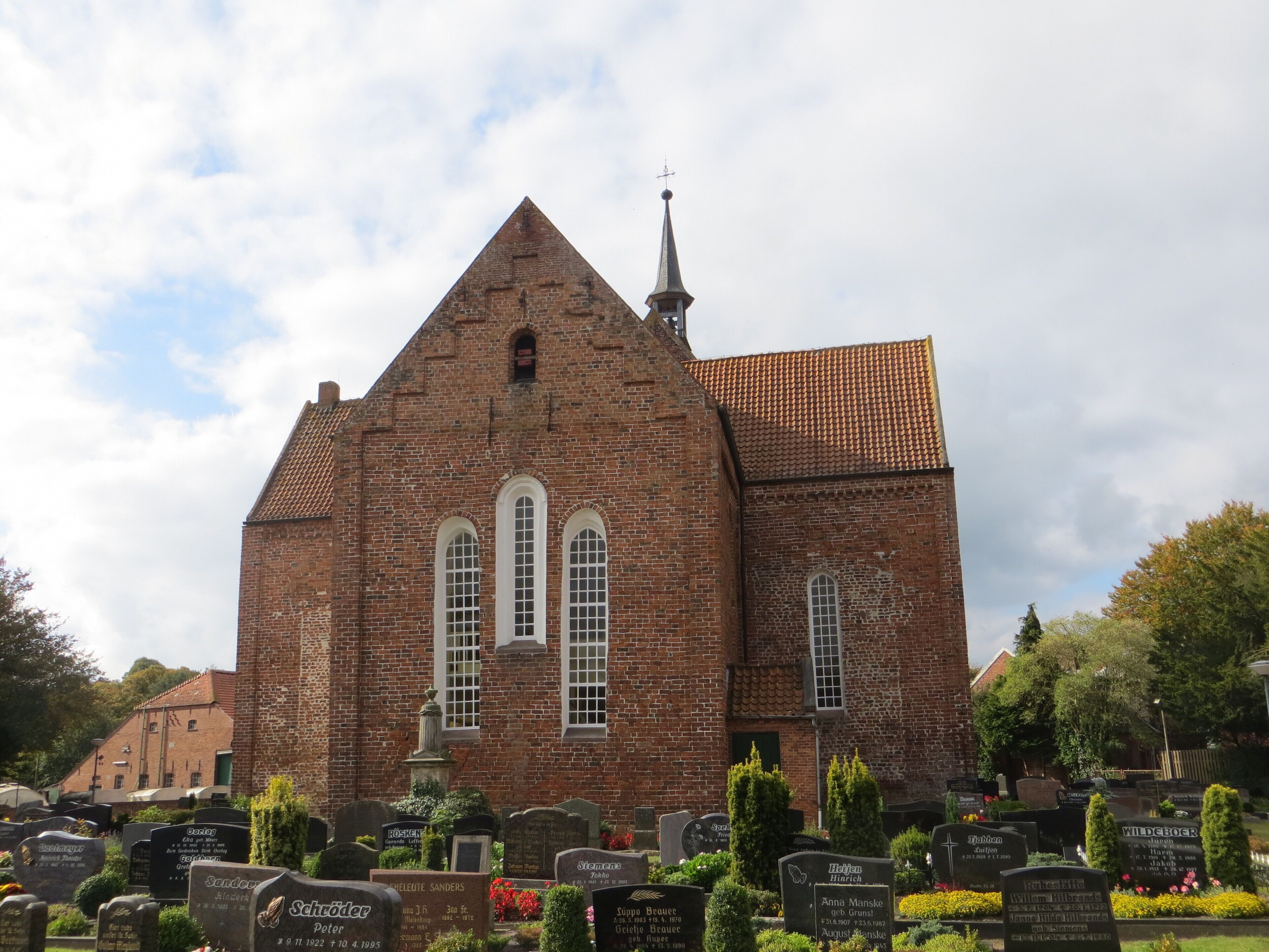 A church with a red brick facade and a tiled roof. In front of the church, there are gravestones with inscriptions.