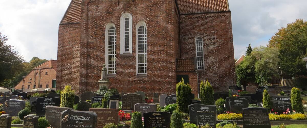 A church with a red brick facade and a tiled roof. In front of the church, there are gravestones with inscriptions.