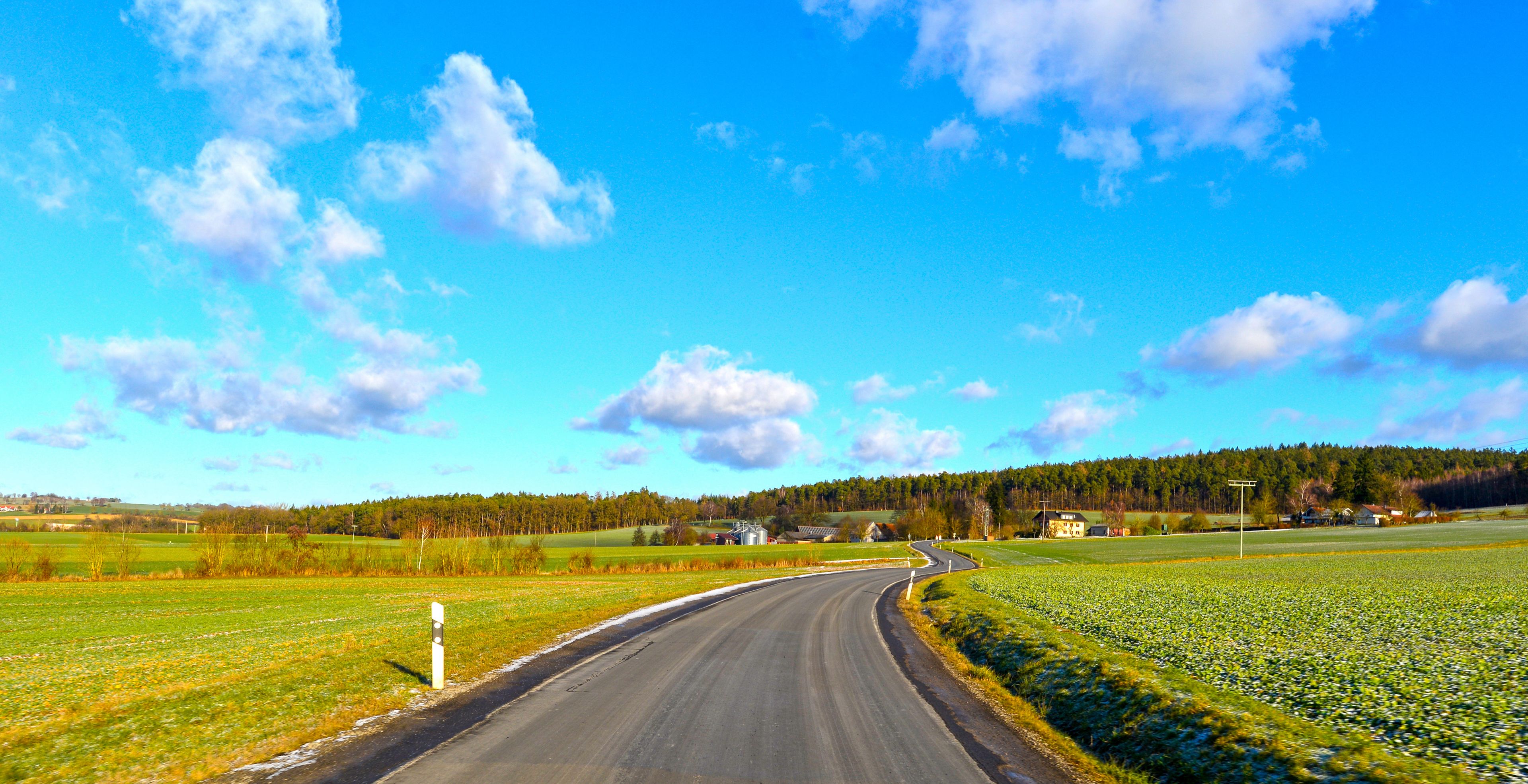 A curving country road with fields on both sides, a forested hill in the distance, and a clear blue sky.