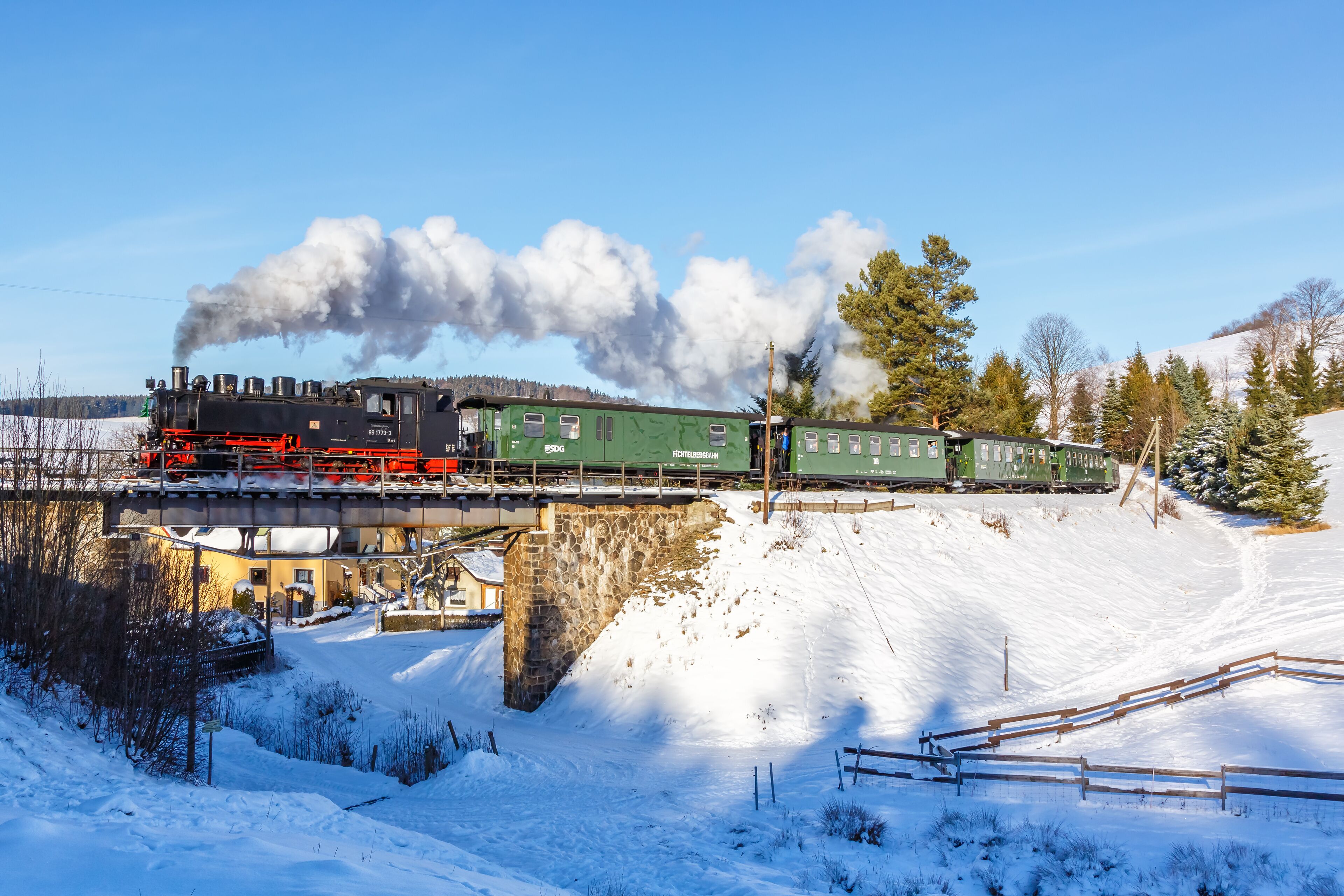 A steam locomotive pulling green passenger cars over a stone bridge in a snowy landscape.