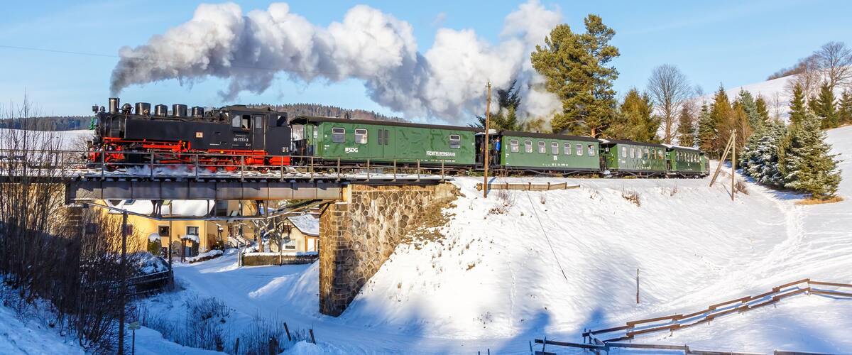 A steam locomotive pulling green passenger cars over a stone bridge in a snowy landscape.